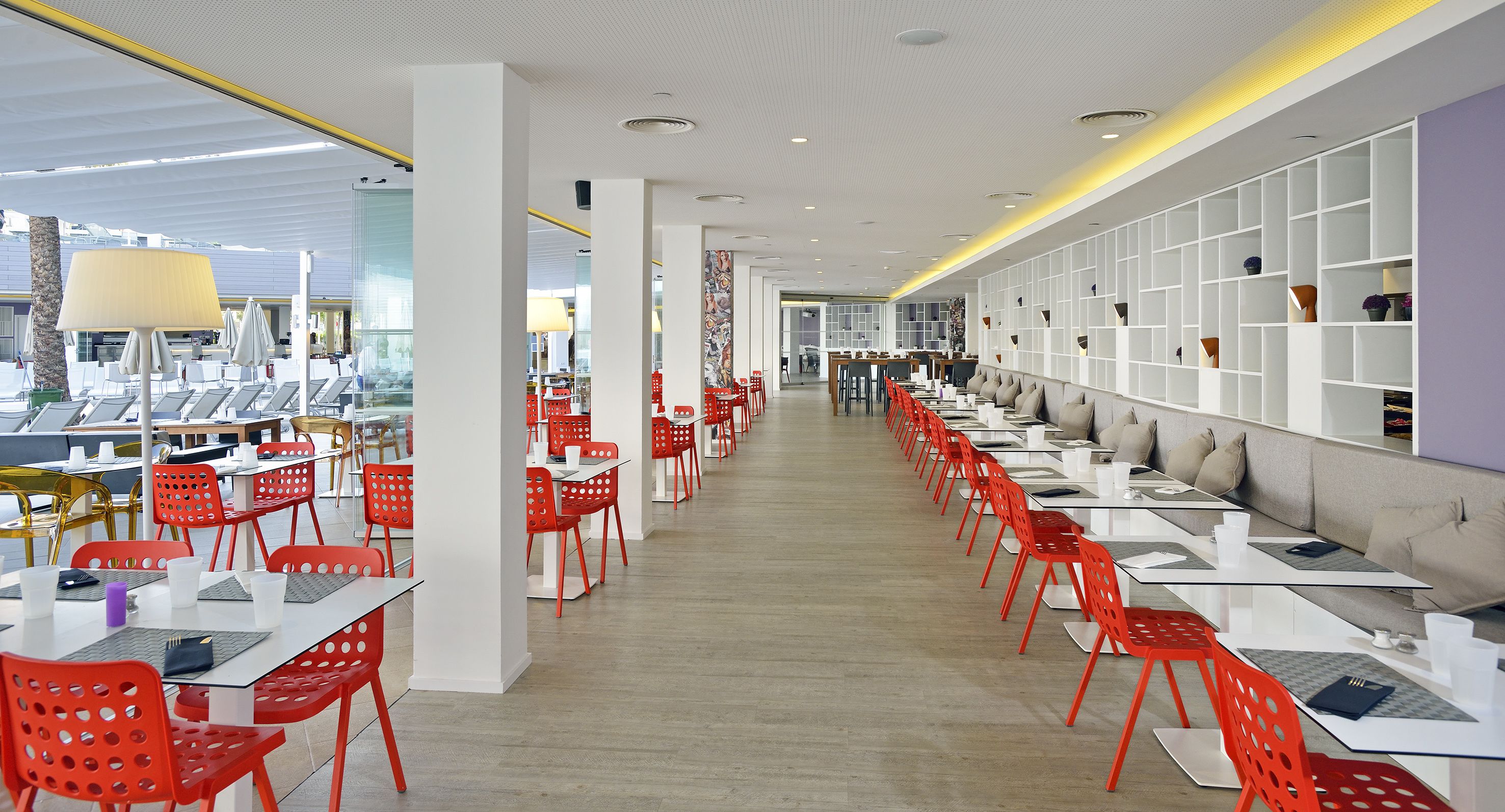 Ein modernes Restaurant mit roten Stühlen und weißen Tischen, einer langen Theke und einer Wand mit Regalen.