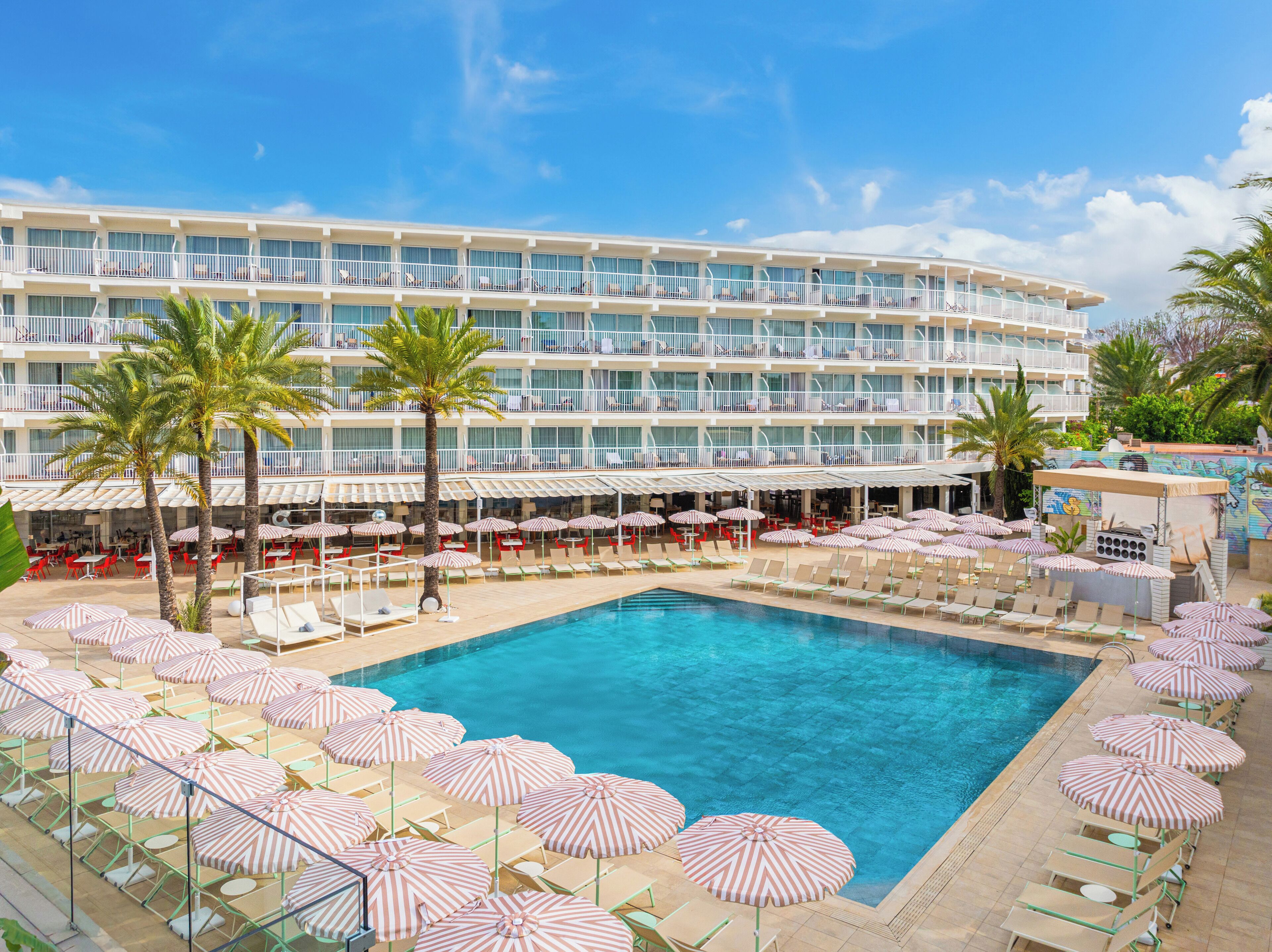 A hotel with a large pool surrounded by sun loungers and striped umbrellas, a multi-story building with balconies, and palm trees.