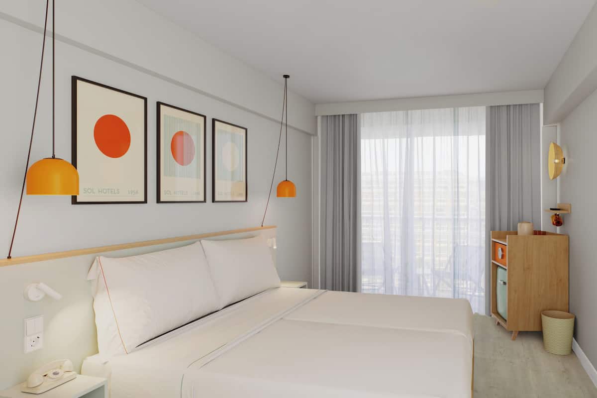 Una habitación de hotel moderna con una cama grande, una mesita de noche con lámpara, un armario y un amplio ventanal con cortinas.
