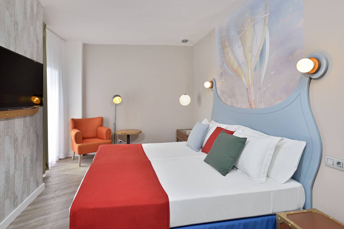 A hotel room with a bed, a chair, a desk, and a TV mounted on the wall.