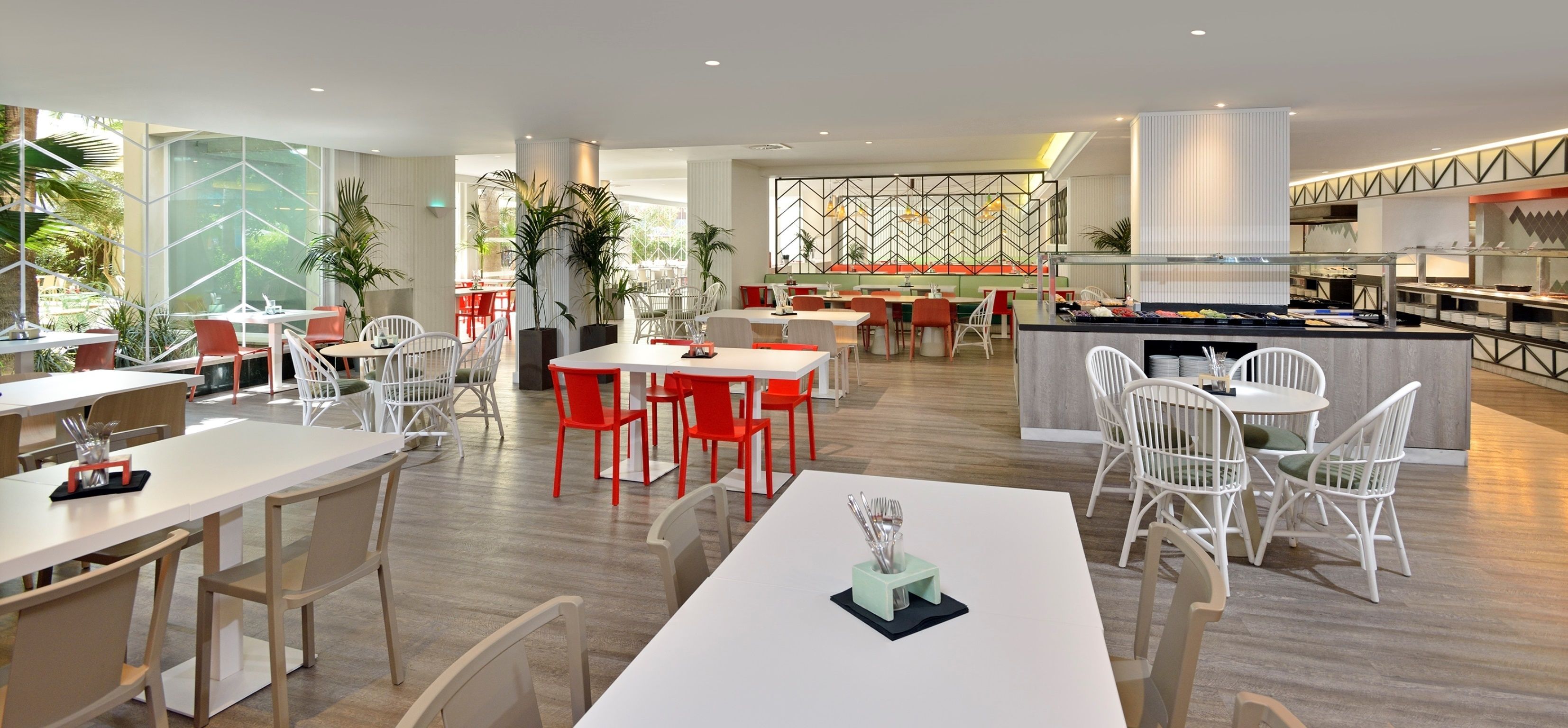 A modern restaurant with white and red chairs, a central dining table, and a kitchen area in the background.