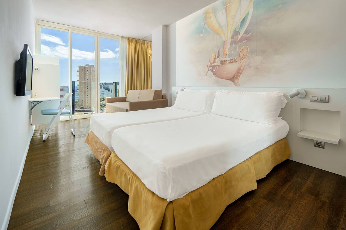 A hotel room with a large bed, a desk, a chair, and a balcony with a city view.