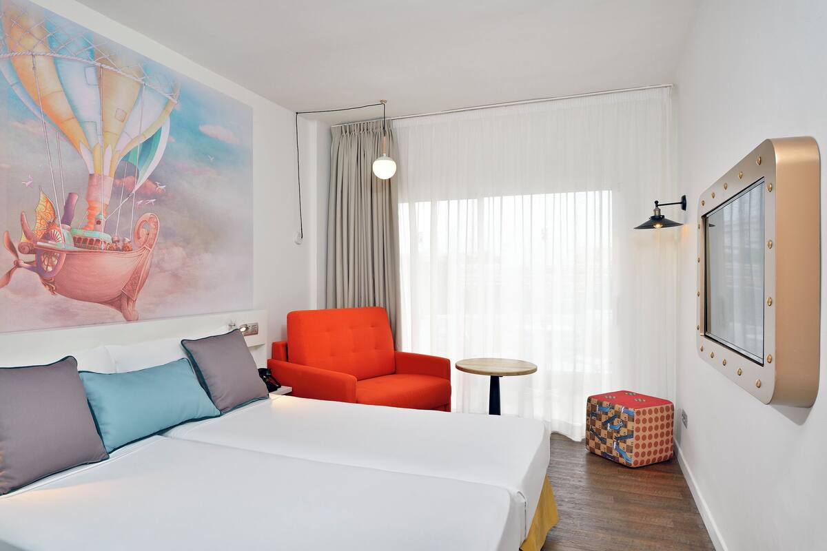A hotel room with a bed, a red armchair, a small round table, a colorful box, and a large painting of a hot air balloon.