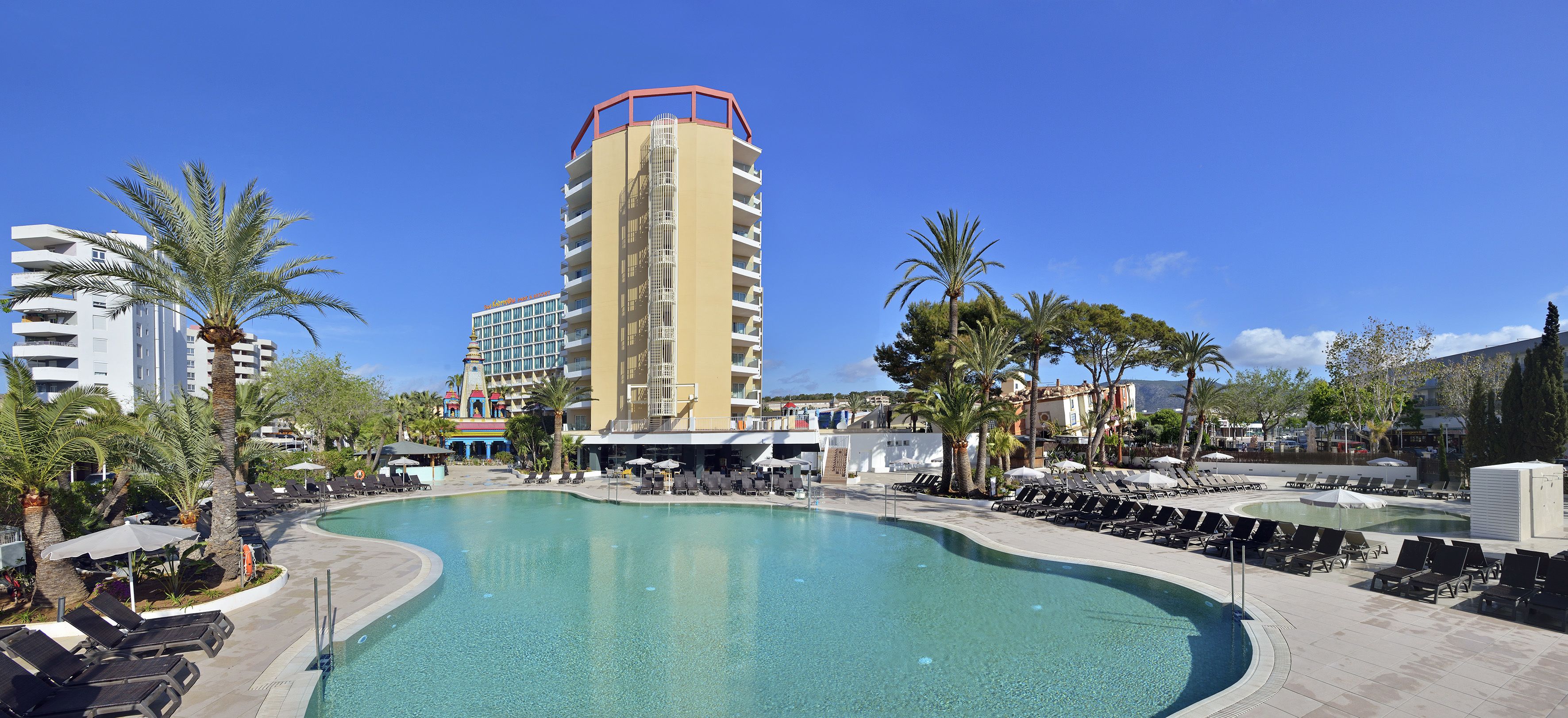 A hotel with a large swimming pool, palm trees, and a tall building with balconies.