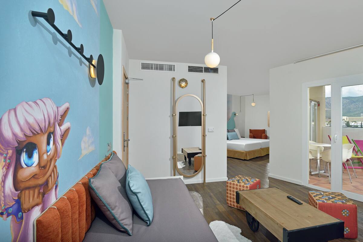 A modern bedroom with a large bed, a sofa, a small table, and a mirror. There is a balcony with a view of the city.