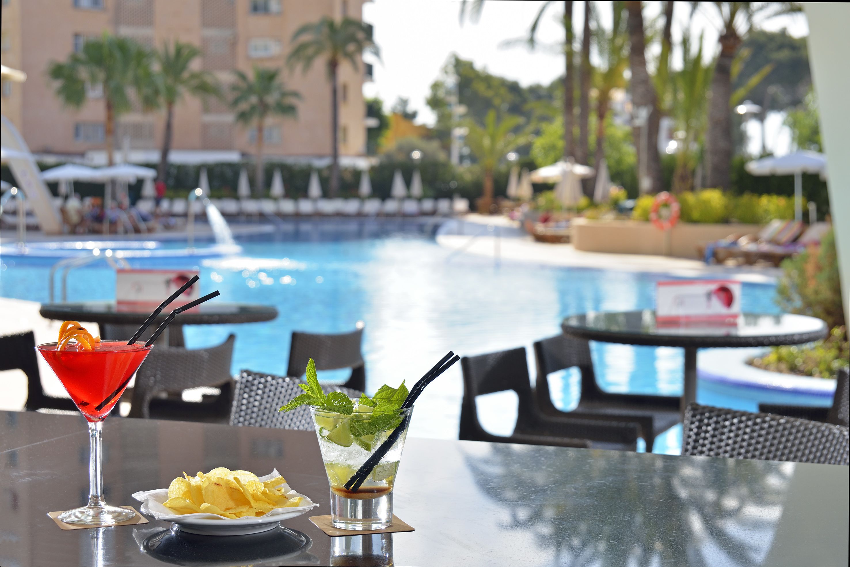 A poolside table with a cocktail and chips, overlooking a pool with lounge chairs and palm trees.