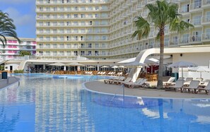 A large outdoor swimming pool with lounge chairs and umbrellas, surrounded by a multi-story hotel building with balconies.