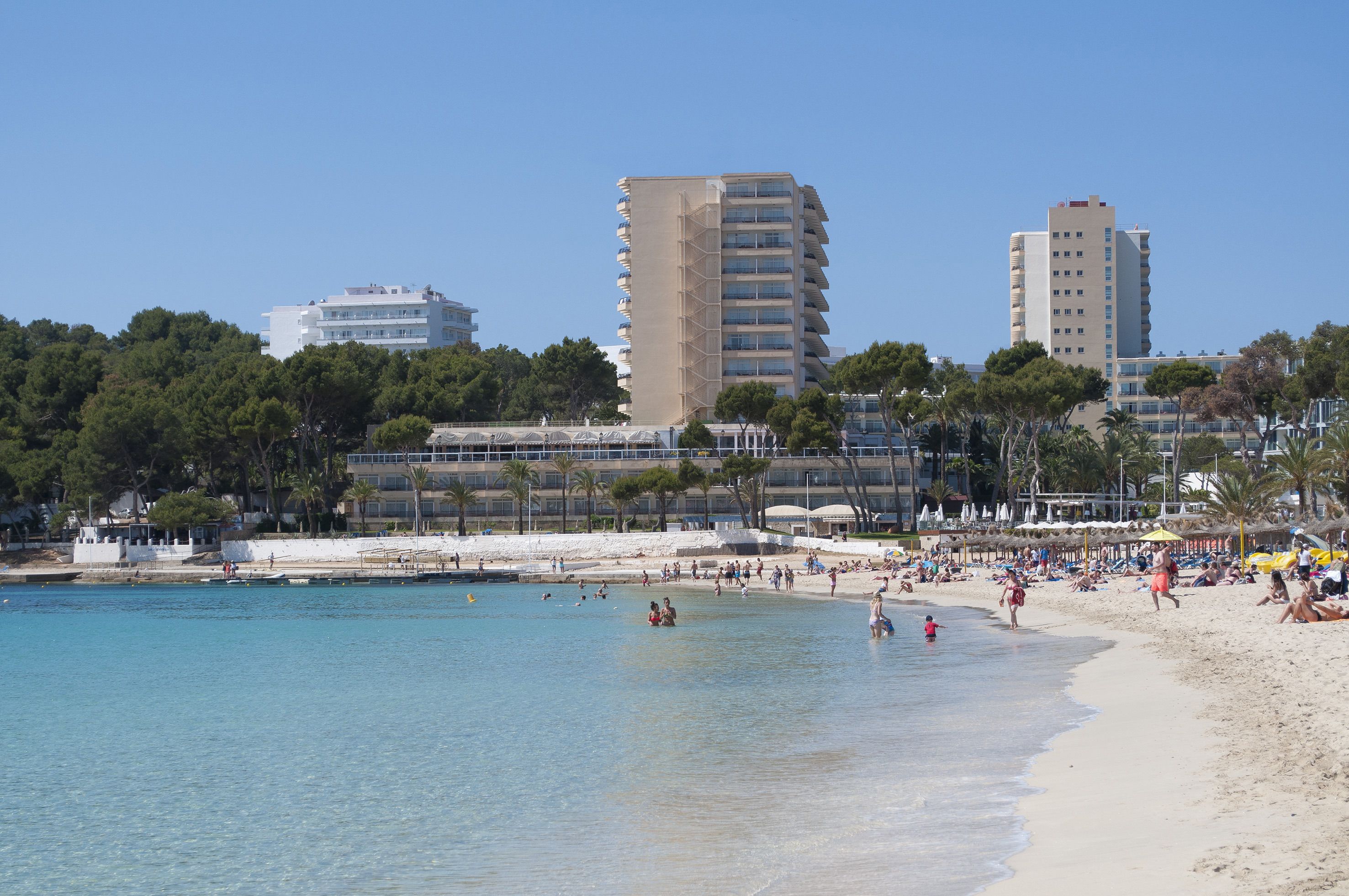 A beach with clear blue water, a sandy shore, and buildings in the background.