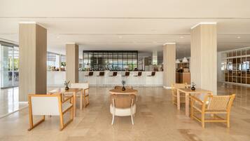 Un lobby moderno de hotel con un área de estar central, un bar y vista a la cocina al fondo.
