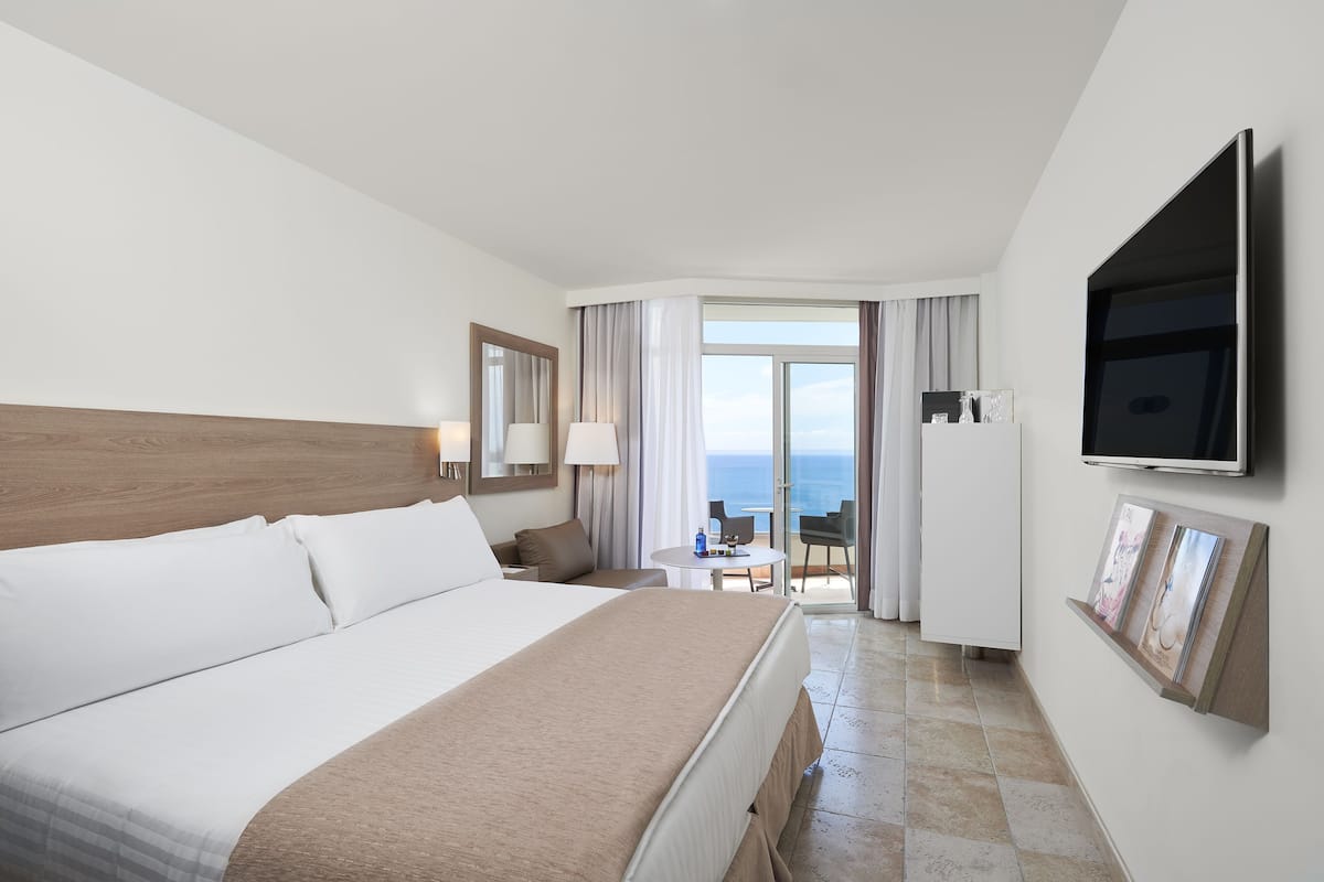 Habitación de hotel con una cama grande, un televisor montado en la pared, una pequeña mesa con una silla y vista al mar a través de un ventanal.