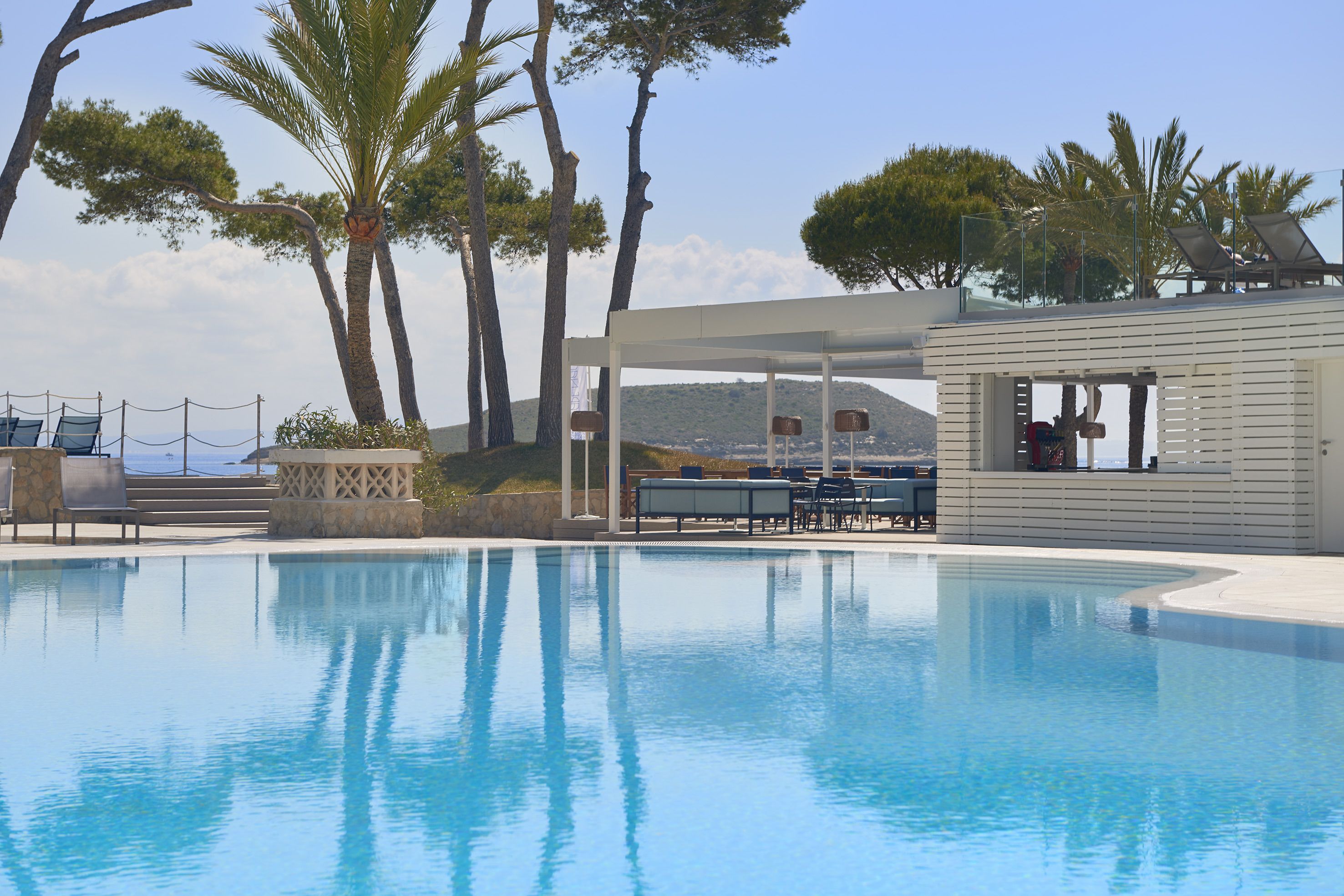 A pool with lounge chairs and a covered seating area, surrounded by trees and a view of the sea.