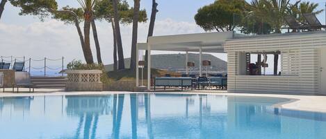 A pool with lounge chairs and a covered seating area, surrounded by trees and a view of the sea.