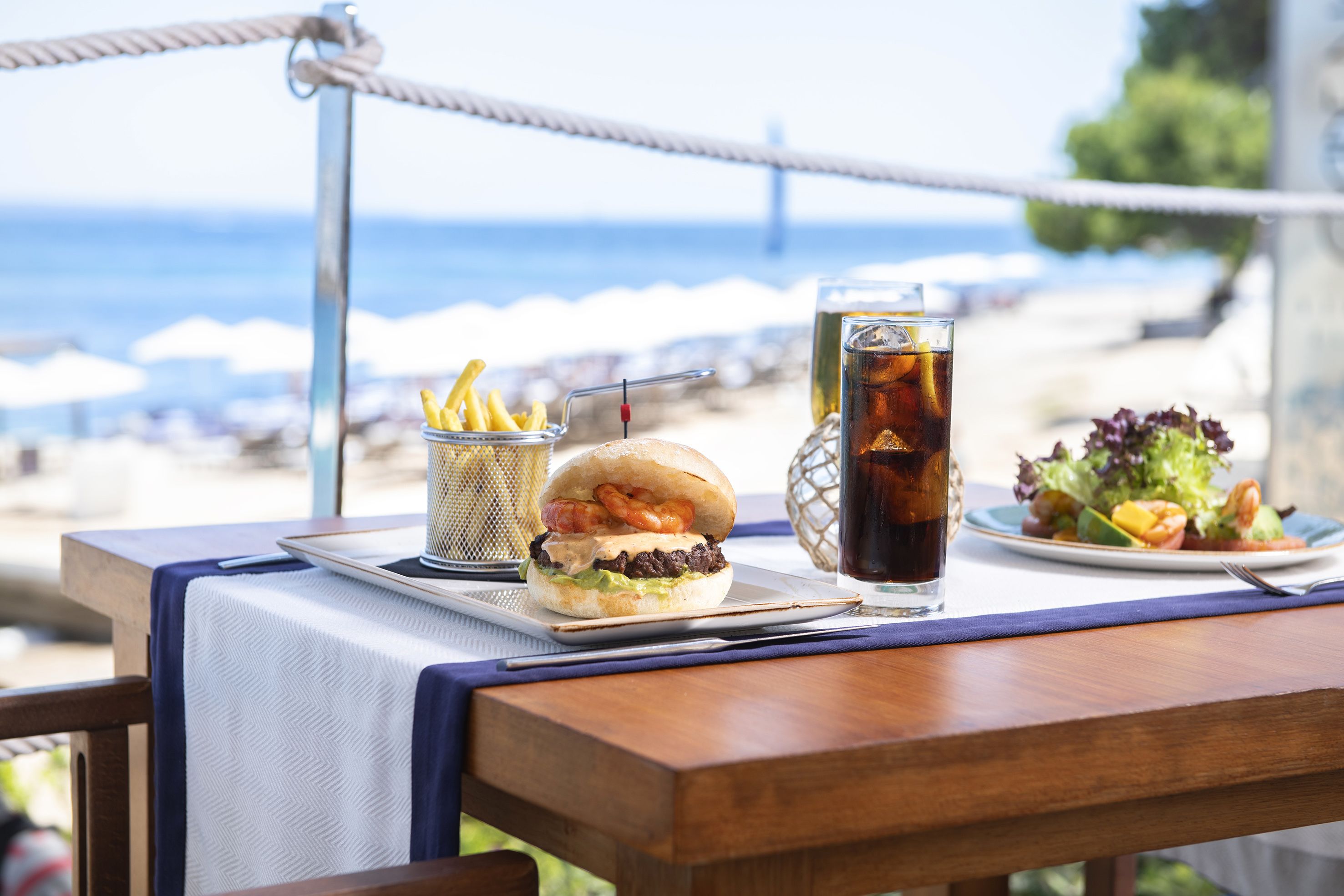 A table with a burger, fries, iced tea, and a salad with shrimp, set against a beach view.