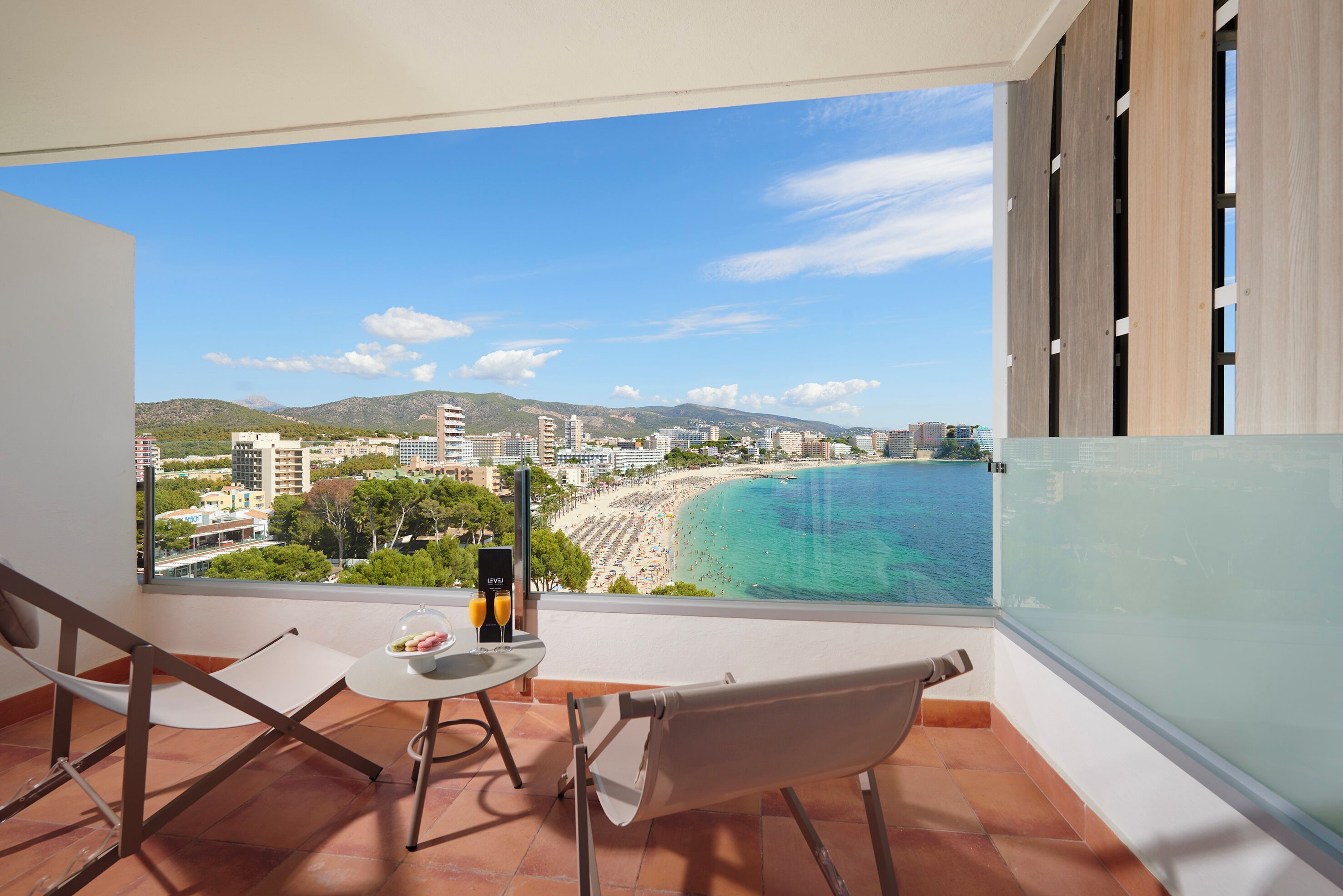 Un balcón con vista a la playa y al paisaje urbano, una mesa con un vaso y una caja, y una silla.