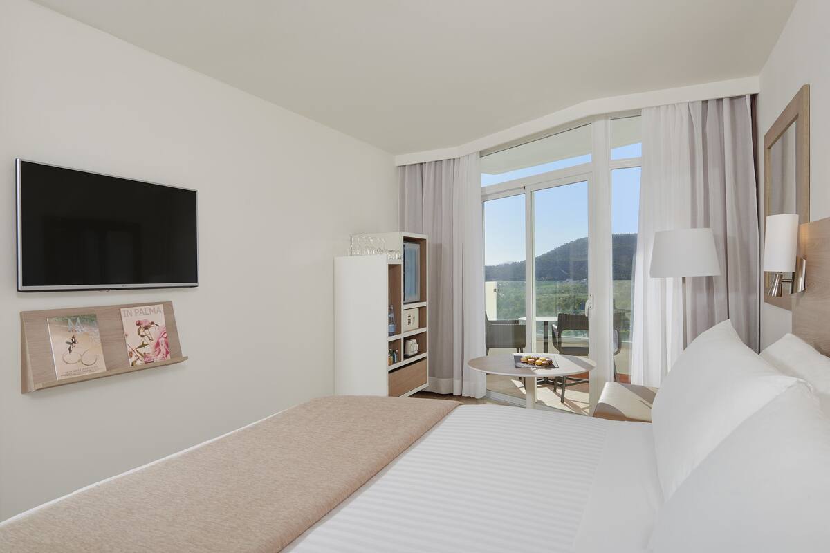 Habitación de hotel con cama, televisor, una mesita de noche con lámpara, estantería y vistas al exterior.