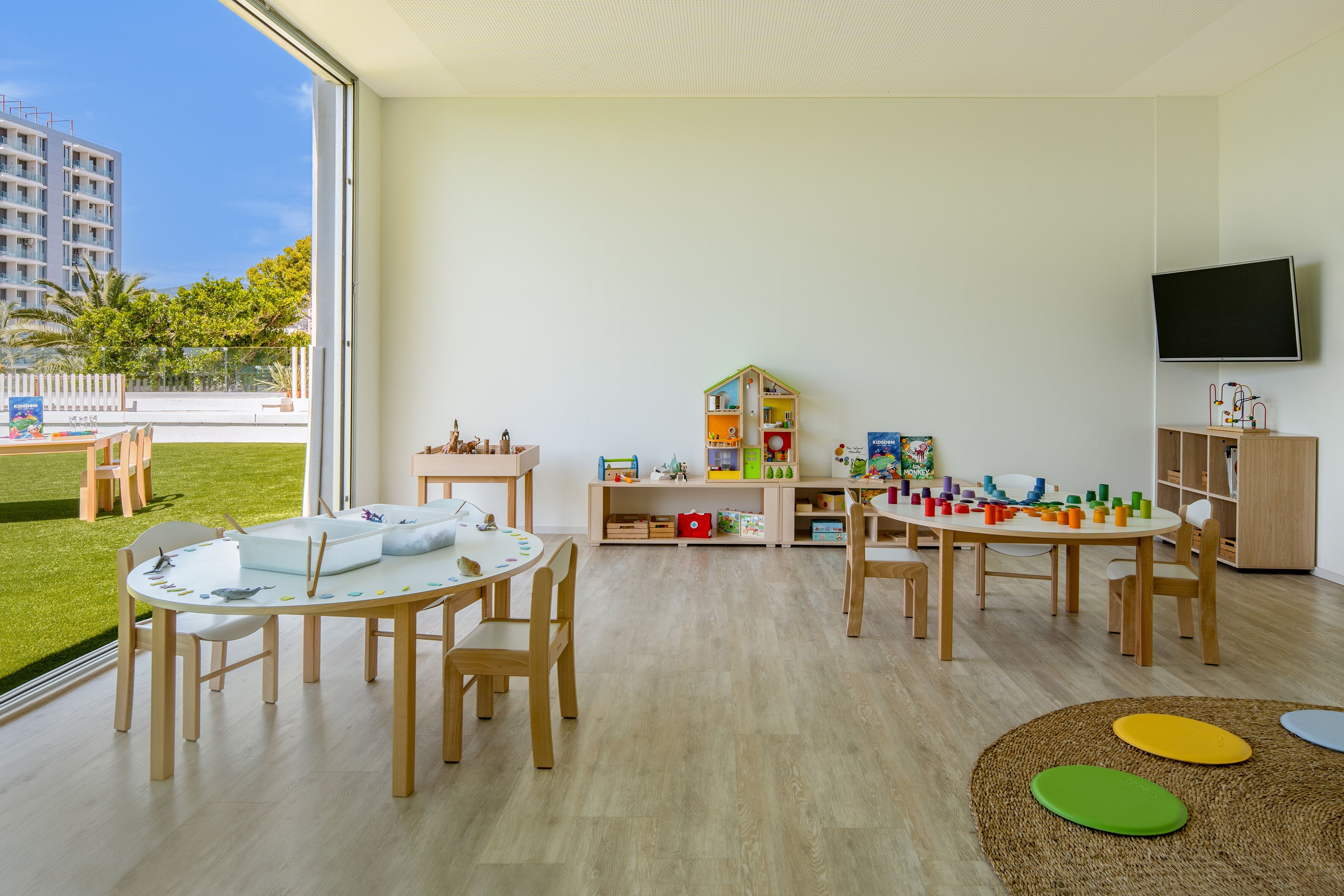 Un salón infantil luminoso y moderno con muebles de madera, un ventanal grande y un televisor montado en la pared.
