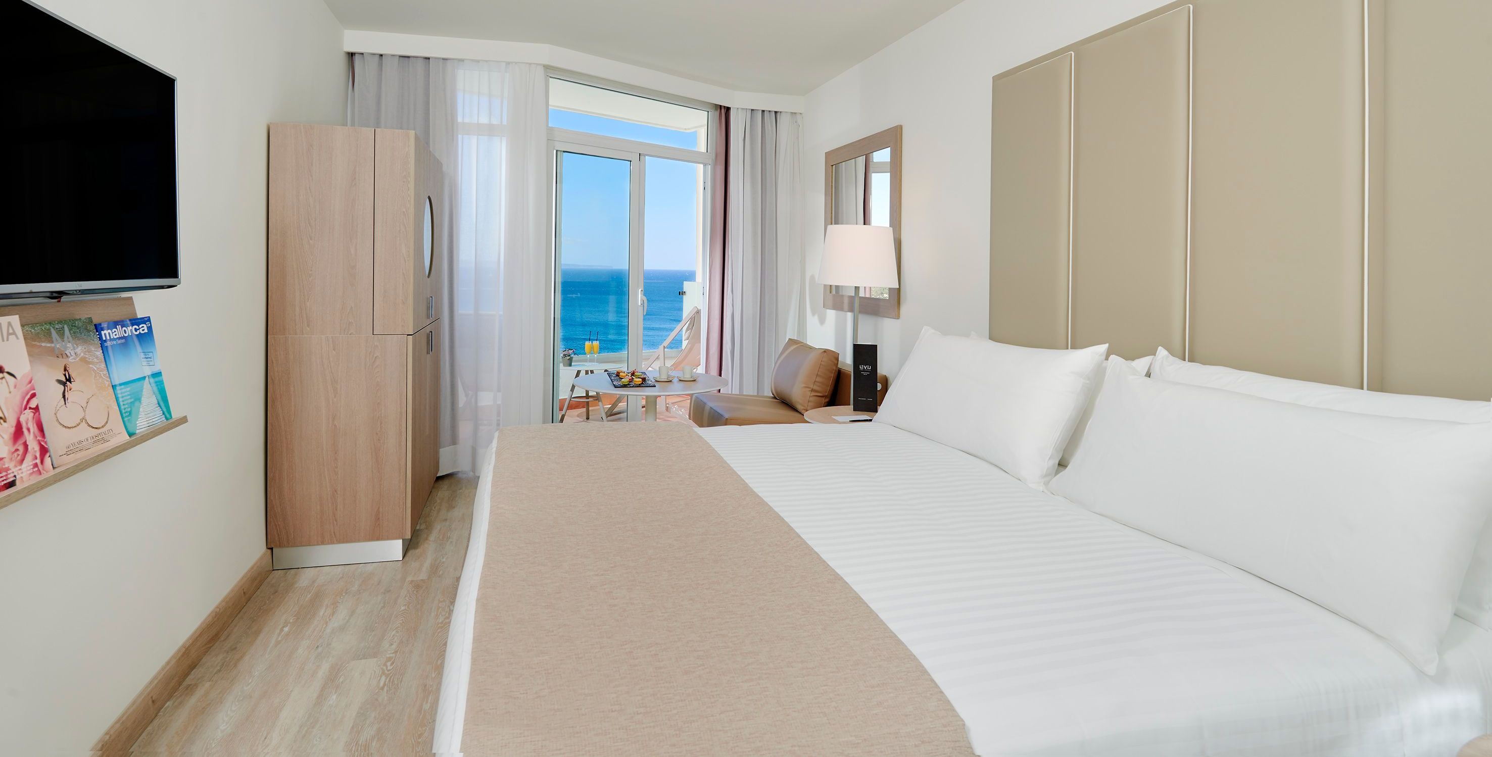 Habitación de hotel con una cama grande, televisión, escritorio con silla y vista al mar a través de un ventanal.