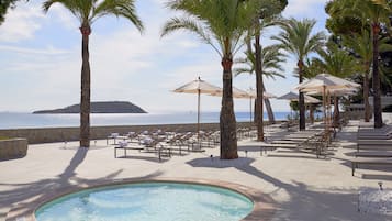 Zona de piscina con sillas de descanso y sombrillas, palmeras y vista al mar.