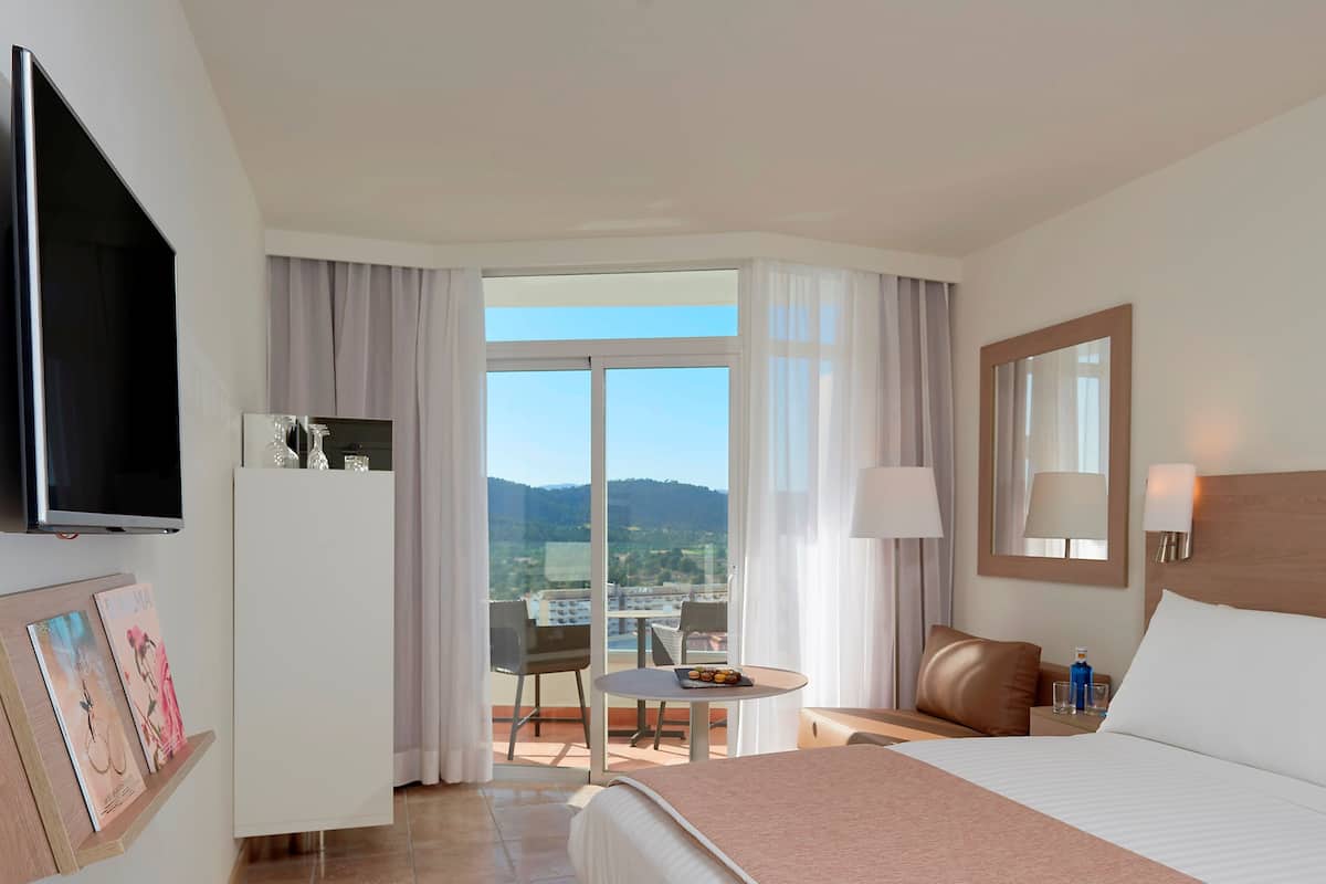 Habitación de hotel con cama, televisión, balcón con mesa y sillas, y vistas al paisaje.