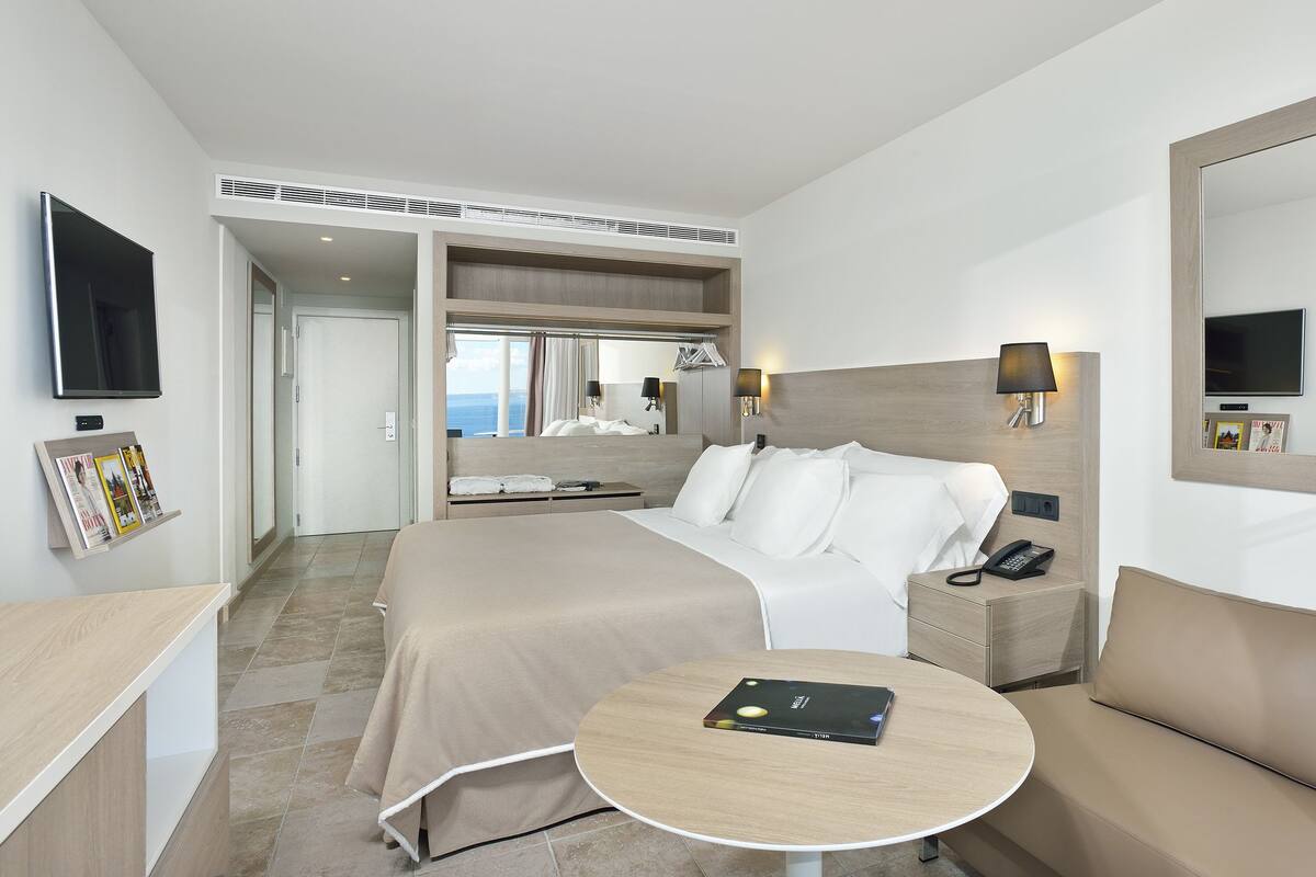 Una habitación de hotel moderna con una cama, un sofá, una mesita, un televisor y un espejo.