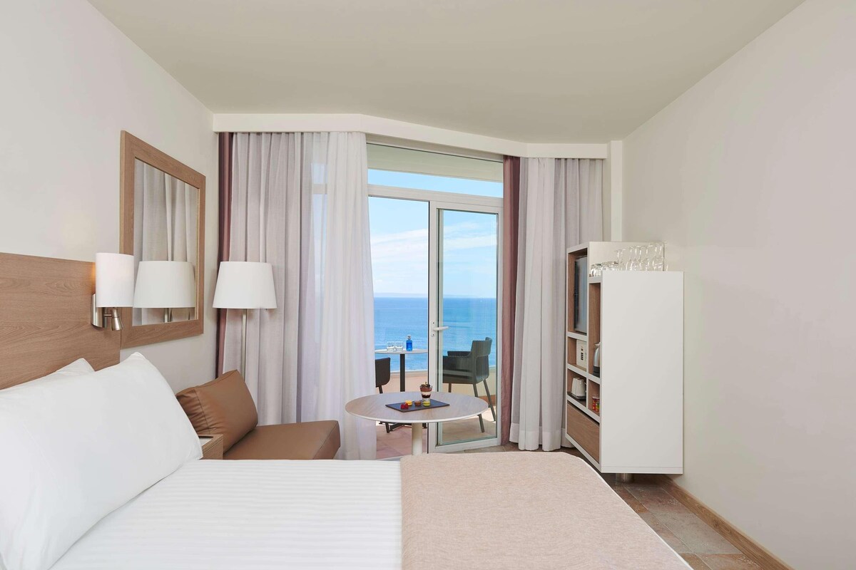 Una habitación de hotel con cama, una mesita con una silla, un minibar y vistas al mar.