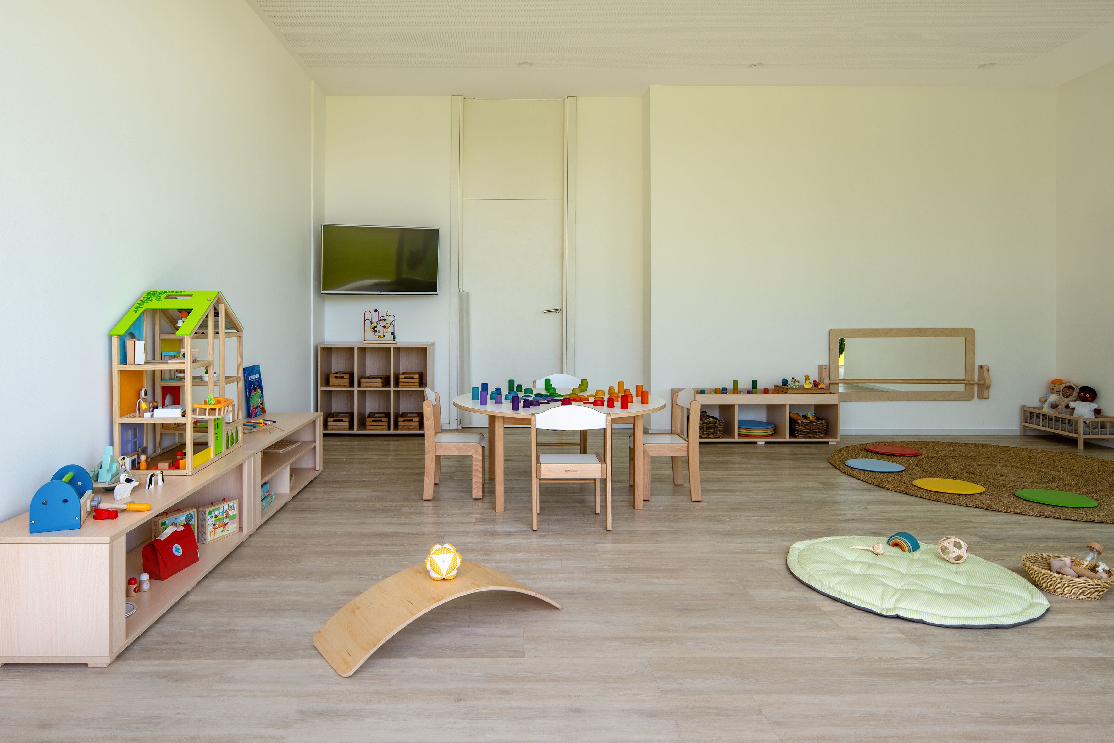 A spacious room with a wooden play area, a television mounted on the wall, and a large window.