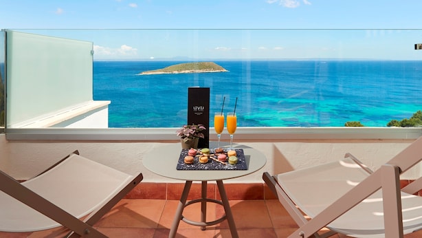 Un balcón con una mesa dispuesta con macarons y dos bebidas, con vista al mar turquesa y una pequeña isla.