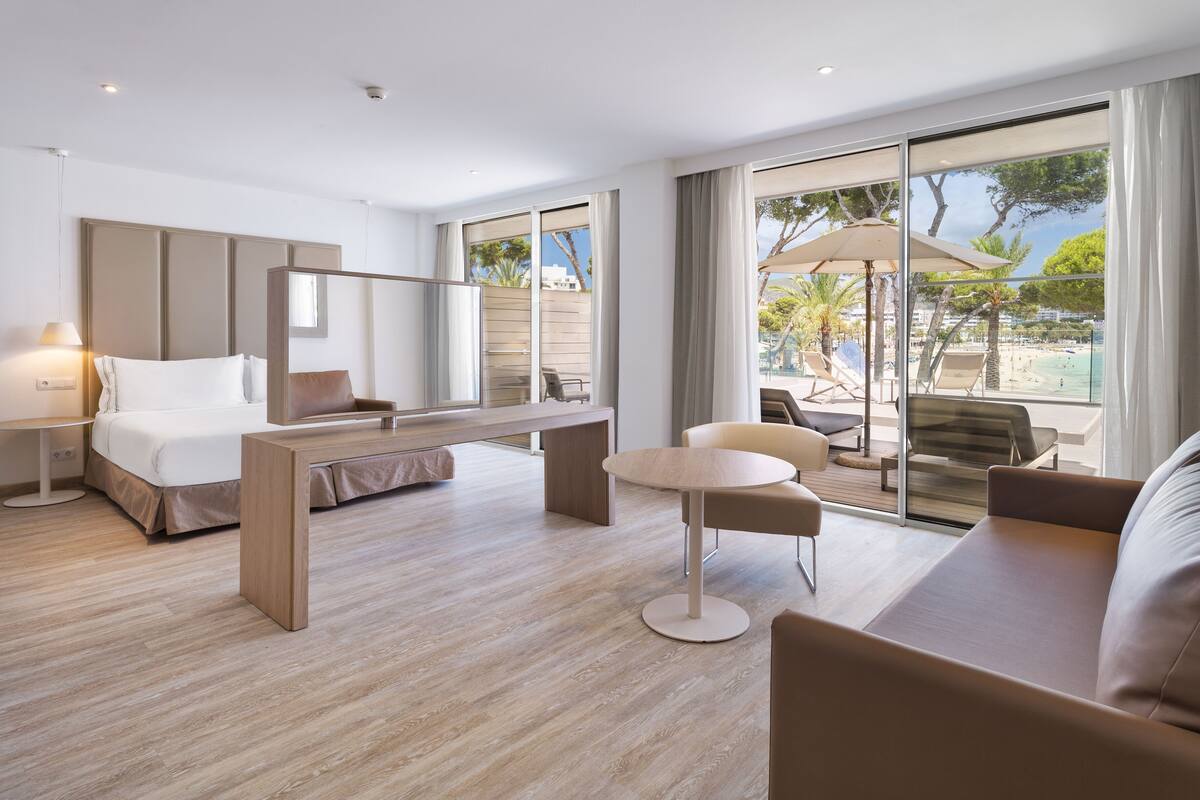 Habitación de hotel moderna con cama, escritorio, mesa redonda y sofá. Amplia ventana con vista a la playa y palmeras.