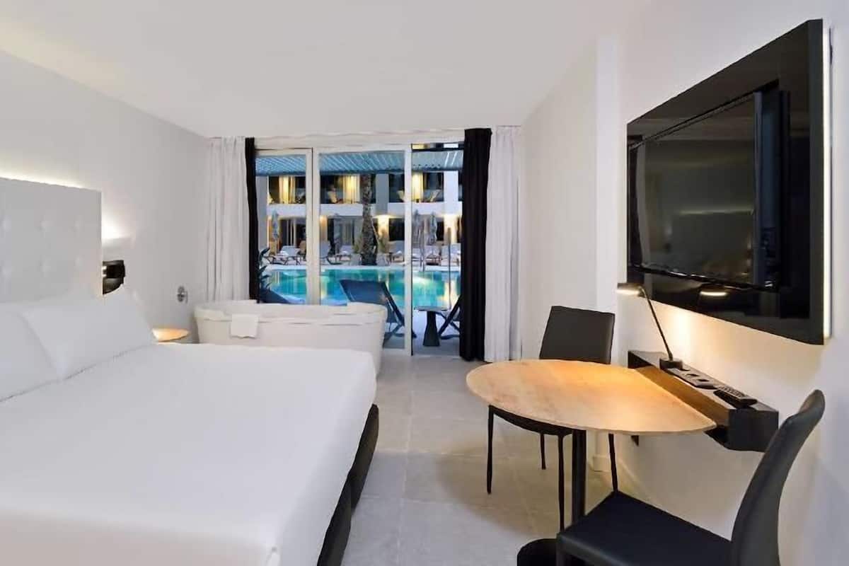 Quarto de hotel com uma cama grande, uma mesa com cadeira, televisão e vista para a área da piscina.