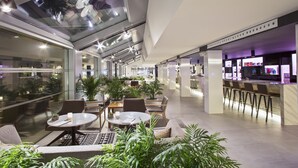 A modern hotel lobby with a glass ceiling, indoor plants, seating areas, and a bar with televisions.