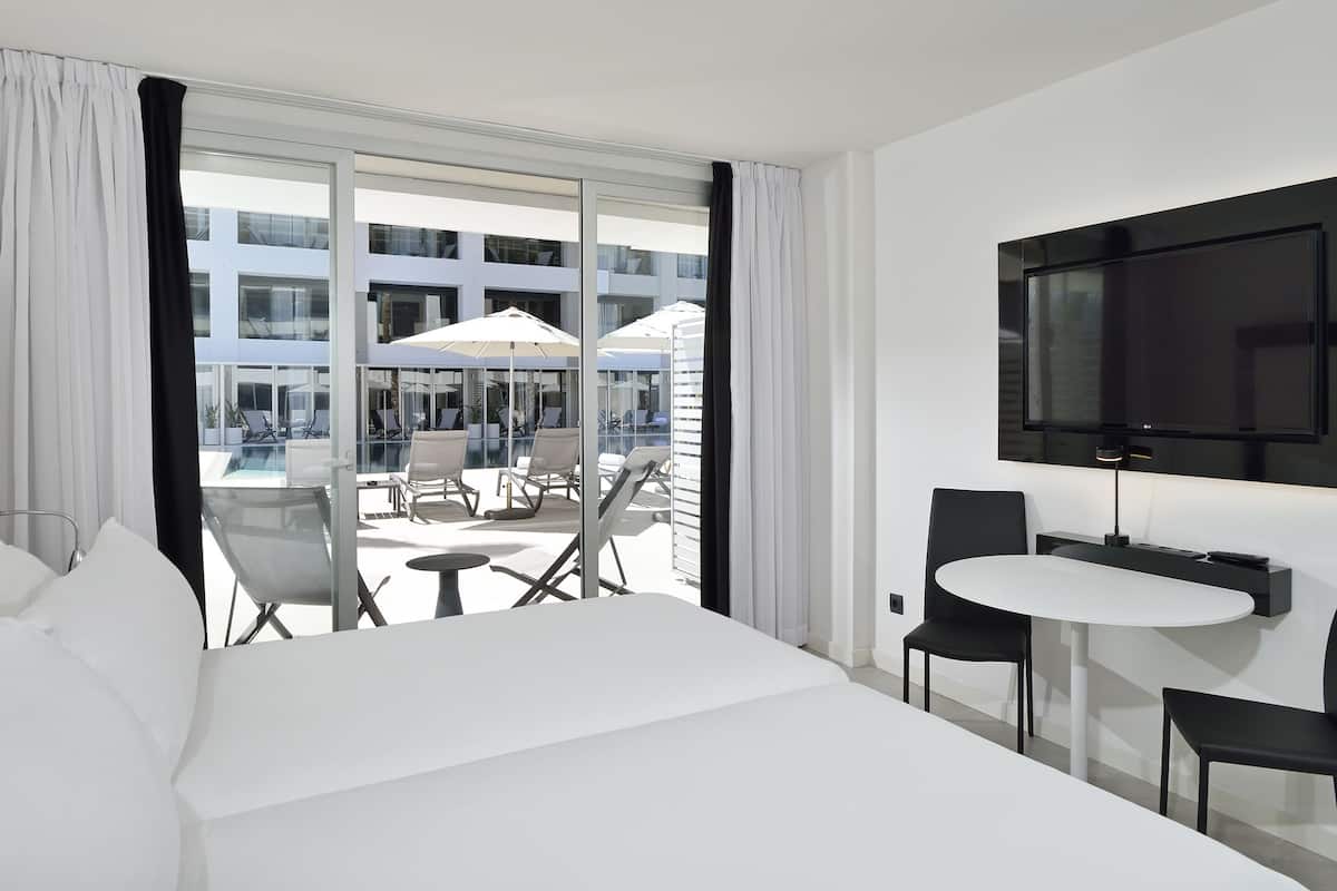 Quarto de hotel com uma cama grande, TV fixada na parede, uma pequena mesa com duas cadeiras e varanda com mobiliário de exterior e piscina.
