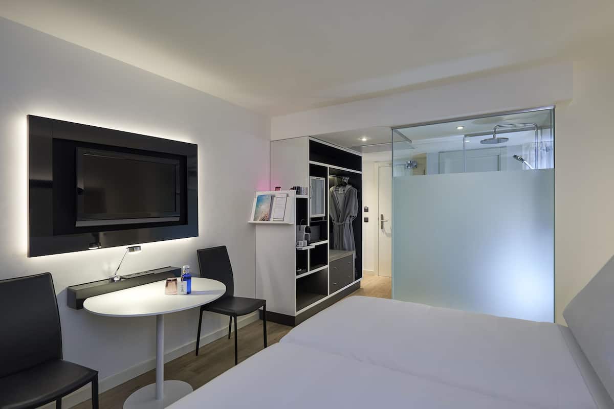 Quarto de hotel moderno com cama, TV fixada na parede, mesa pequena com cadeiras, armário com espelho e área de duche.