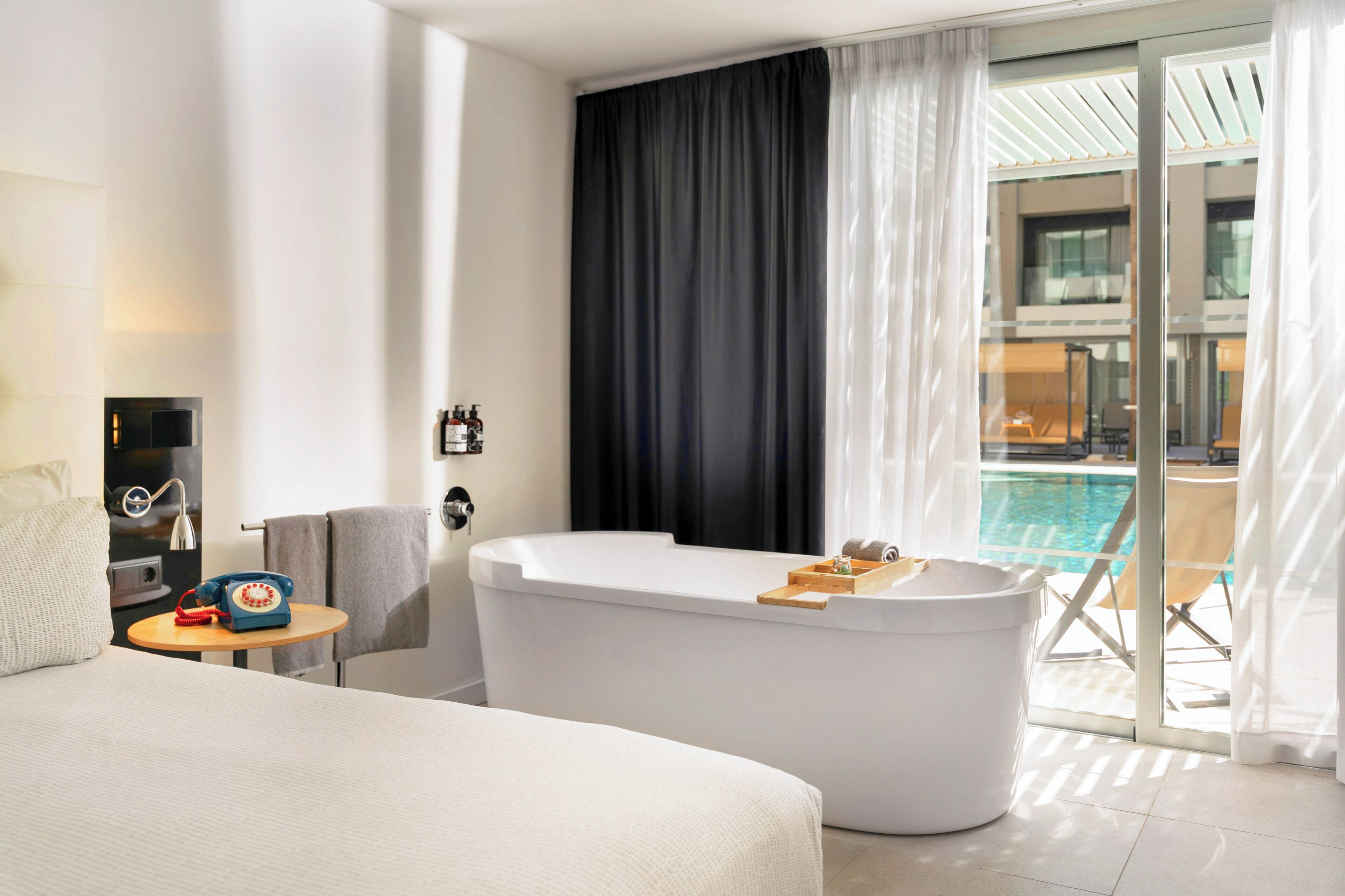 A modern hotel room with a large bed, a bathtub, a small table with a telephone, and a view of a swimming pool.