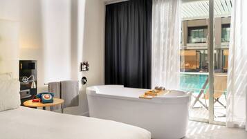 A modern hotel room with a large bed, a bathtub, a small table with a telephone, and a view of a swimming pool.