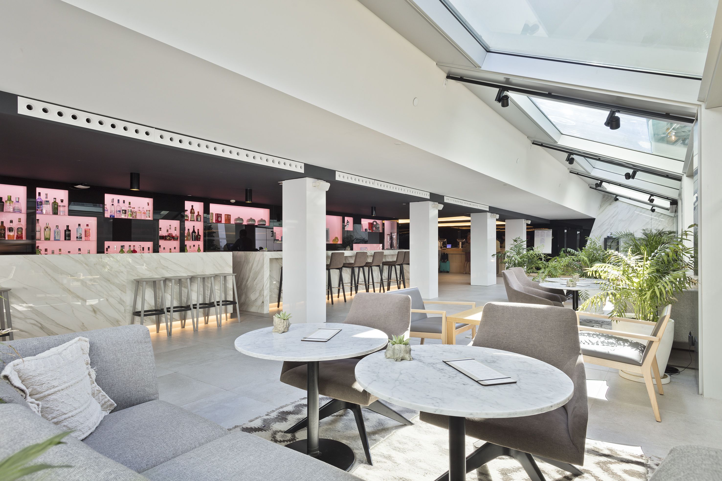 Un amplio espacio interior con mesas redondas y asientos cómodos, una zona de bar moderna al fondo y un techo con tragaluces.