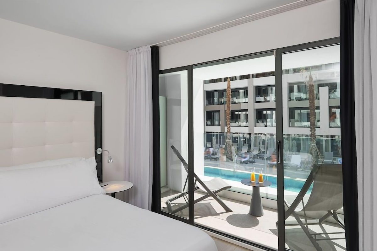 A hotel room with a large window, a bed, a small table, and a view of a pool and building.