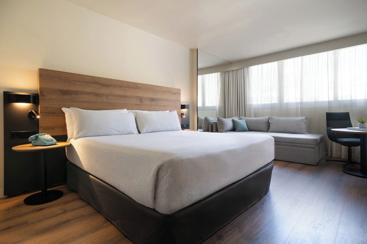 Quarto de hotel moderno com uma cama grande, mesas de cabeceira, um espaço de estar e vista para o exterior.