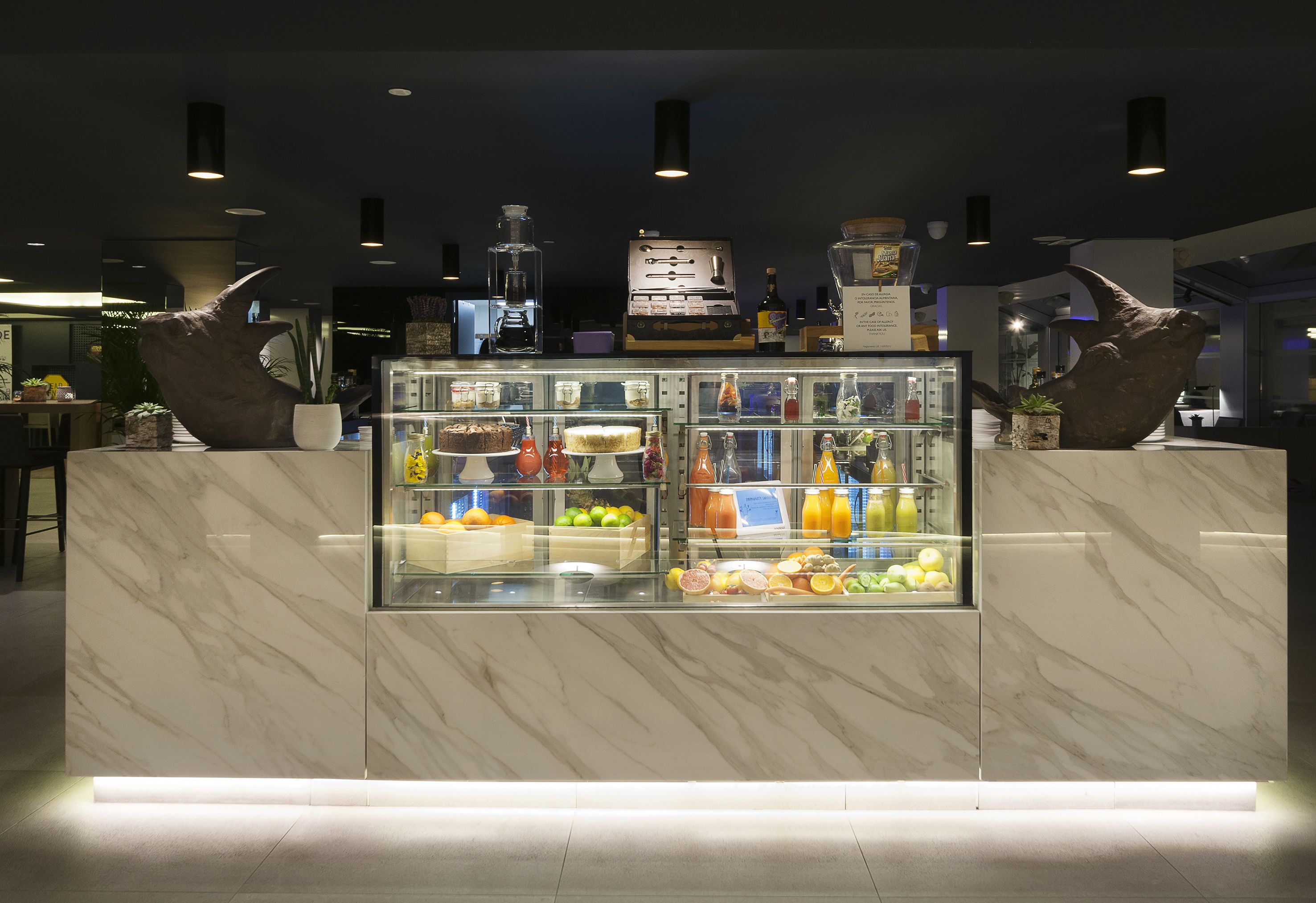A marble counter with a refrigerated display case filled with fruits and beverages, a coffee machine, and decorative items.