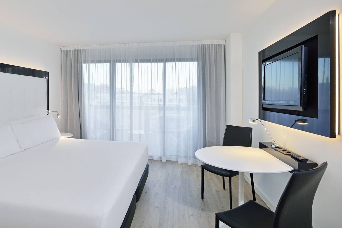 Quarto de hotel com uma cama grande, uma mesa redonda pequena, uma TV fixada na parede e vista da cidade através de cortinas translúcidas.