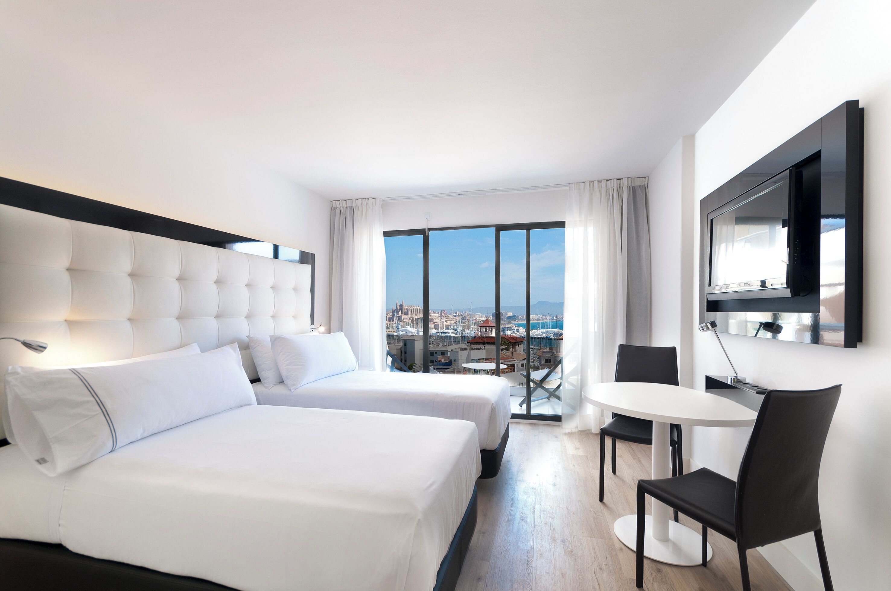 A hotel room with two beds, a TV, a dining table with chairs, and a view of a cityscape.