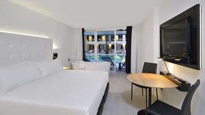 A modern hotel room with a large bed, a dining area, and a view of an outdoor pool.