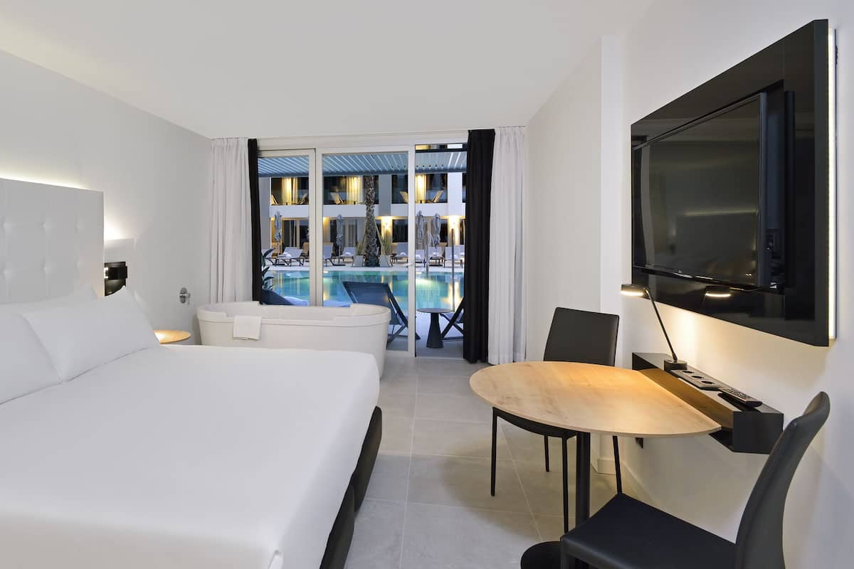 Quarto de hotel moderno com uma cama grande, área de refeições e vista para uma piscina ao ar livre.