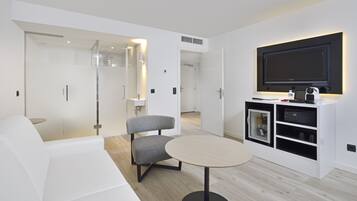 Habitación de hotel moderna con sofá blanco, sillón gris, mesa redonda, minibar y televisión montada en la pared.