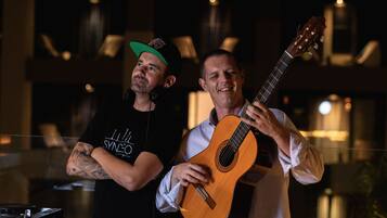 Two men, one playing a guitar and the other DJing, in a dimly lit room with a modern interior design.