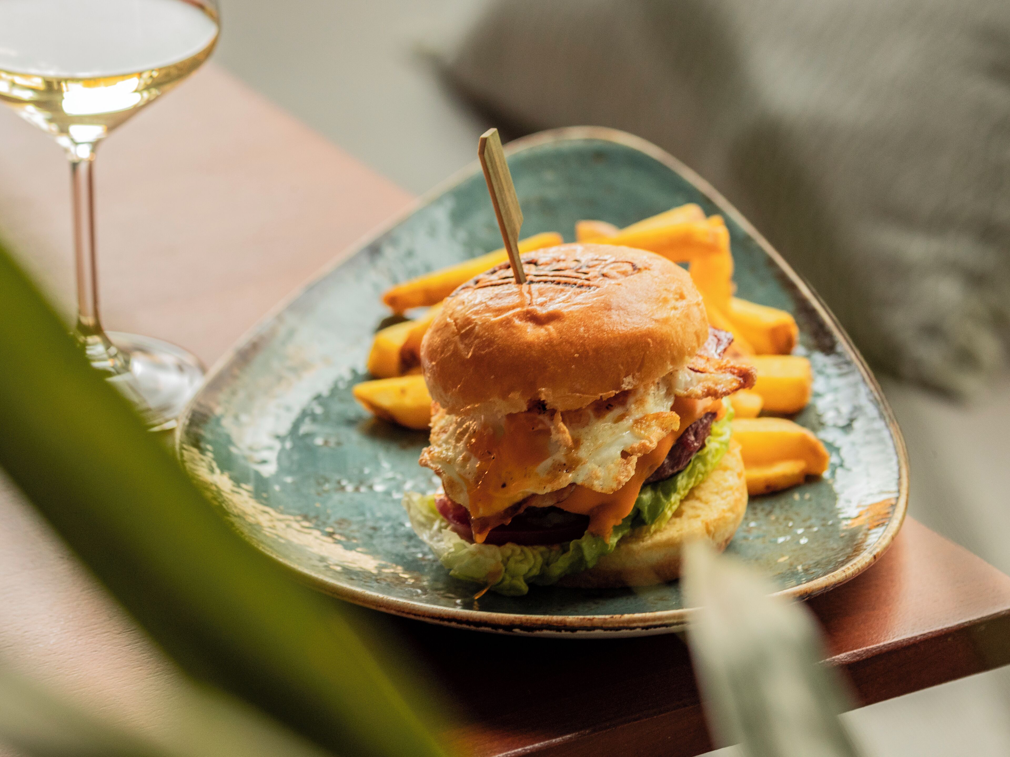 Un plato con una hamburguesa y papas fritas, una copa de vino blanco y una planta verde al frente.