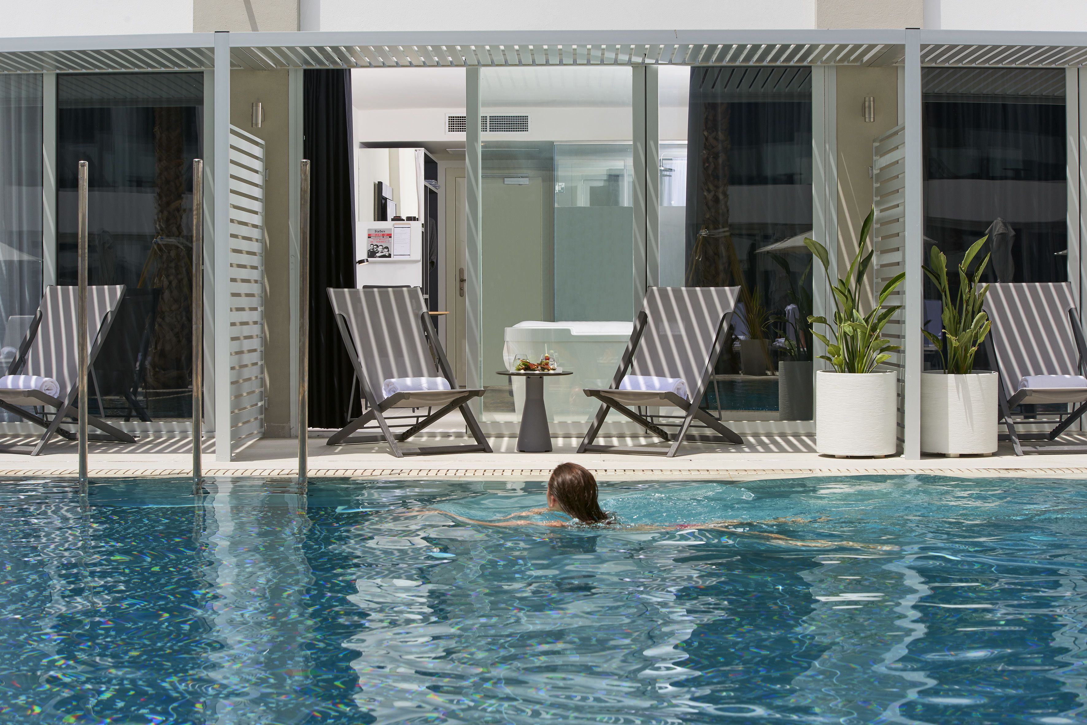A swimming pool with lounge chairs and potted plants, surrounded by glass doors and a modern building.