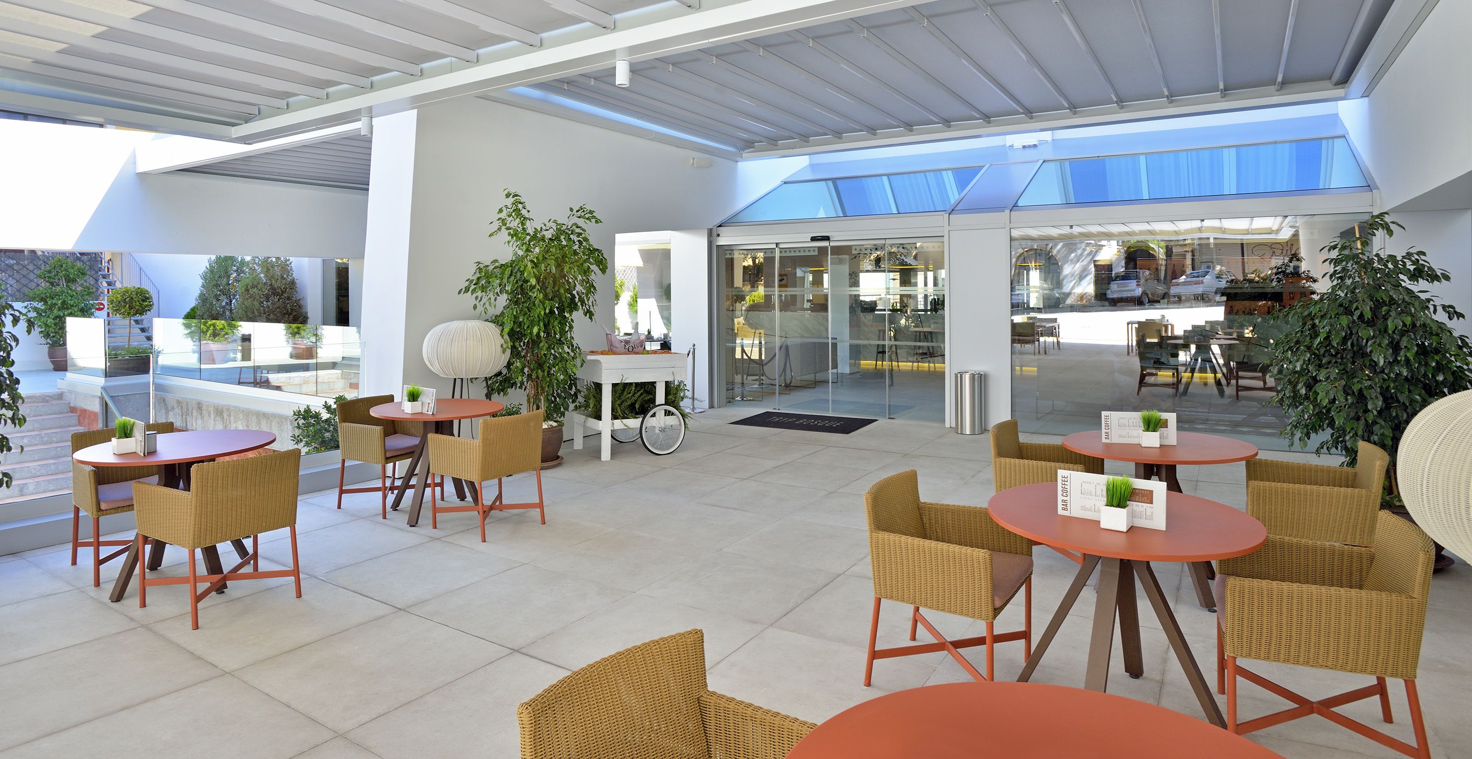 A modern outdoor seating area with wicker chairs and round tables, featuring a glass door entrance and potted plants.