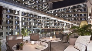A modern hotel atrium with multiple dining tables and chairs, a central staircase, and a large indoor garden area.