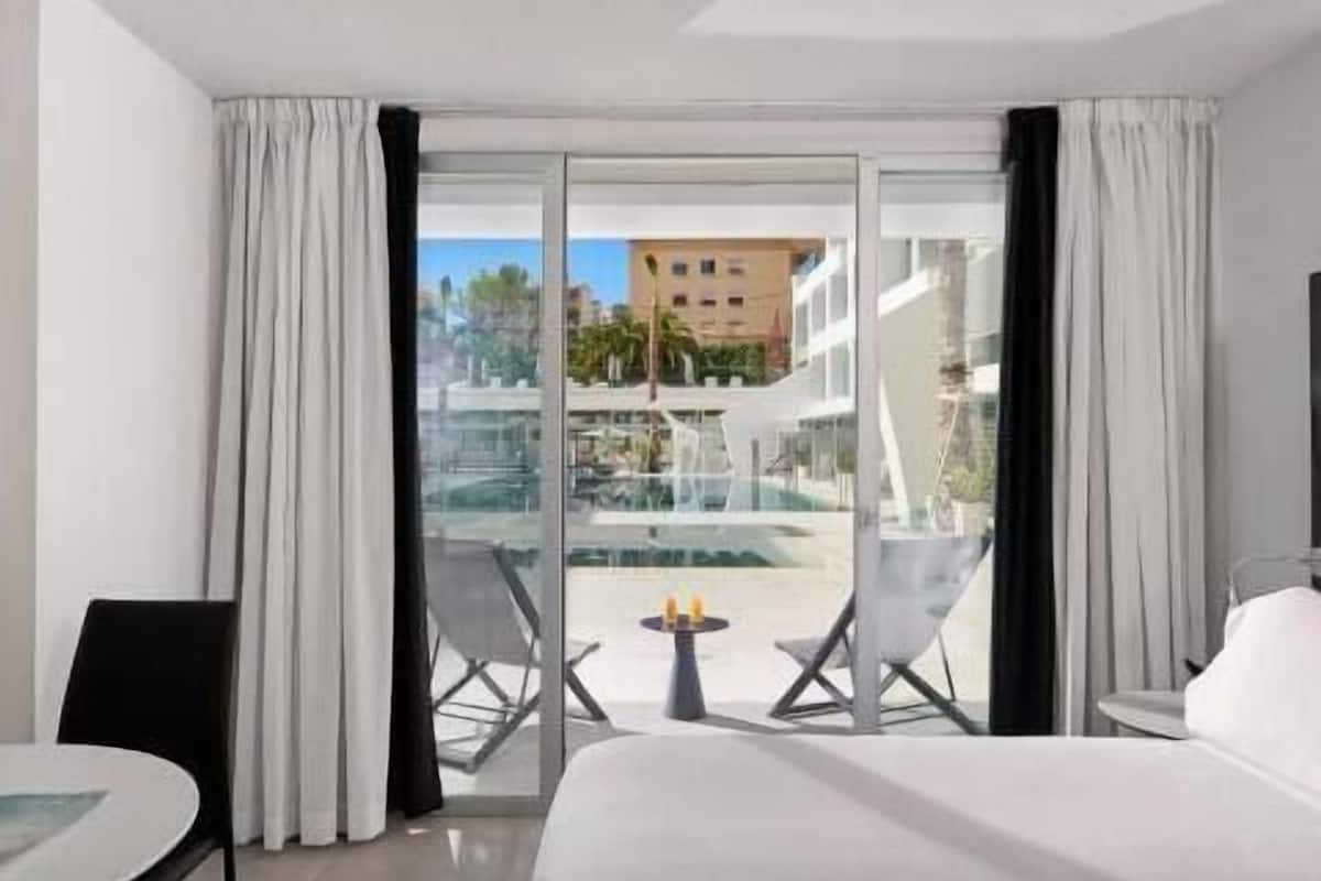 Quarto de hotel com cama, uma mesinha, duas cadeiras e vista para uma piscina e edifícios.