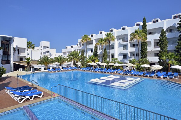 Una piscina con sillas de descanso azules y sombrillas, rodeada de edificios blancos con balcones y palmeras.